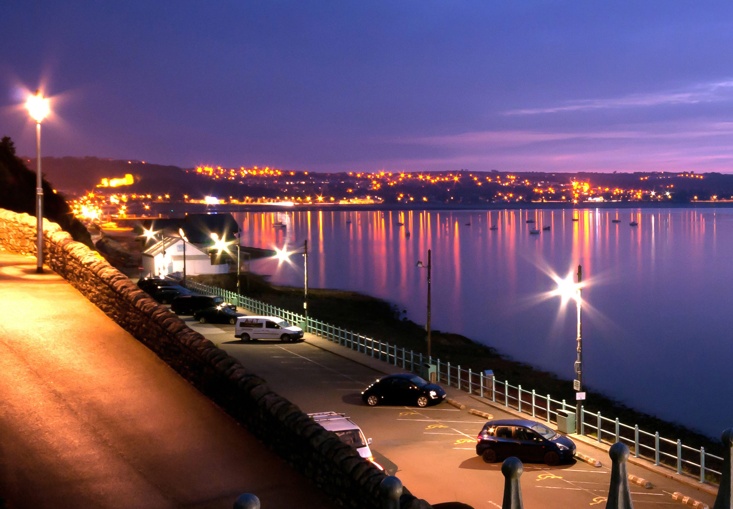 A bay on the south Wales coast at dusk A lit promenade and car park above a bay at dusk, with town lights across the water