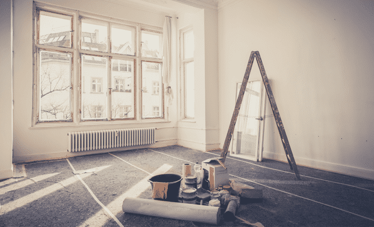 A photo of a living room being refurbished. A ladder stands near some windows and tools and pots of paint are on the floor.