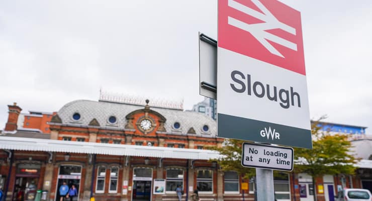 A close up of a sign that says 'Slough' with Slough Train Station in the background