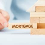 A financial consultant completes a wooden model of a house. The final piece of wood being slotted in has the word 'mortgage' written on it.