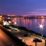 Full tide in Swansea Bay in South Wales. Photo taken at night and features a calm ocean, cars in a car park and the city of Swansea in the background.