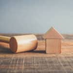 A wooden hammer sits on a surface next to a wooden house. Image represents a property auction.