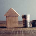 A wooden house sits next to a pile of coins on a table surface. Image represents house value.