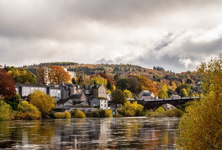 An autumnal scene of Perth in Scotland. The city is seen among red, yellow and orange trees.