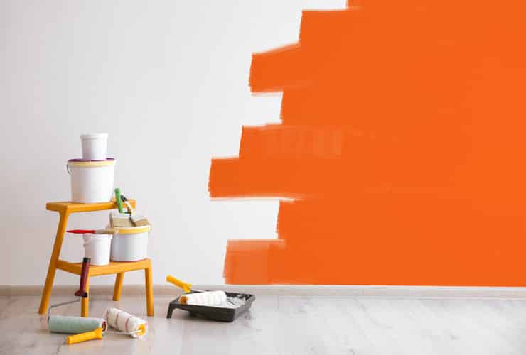 A wall is half painted orange. There is a step ladder with paint pots rested on it.