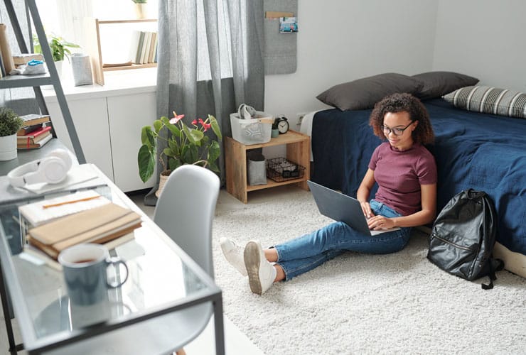 Young contemporary female student in casualwear sitting on the floor by bed with laptop on legs during online lesson in home environment.