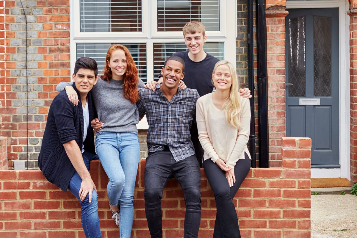 Photograph of a group of smiling college students outside a rented, shared house.