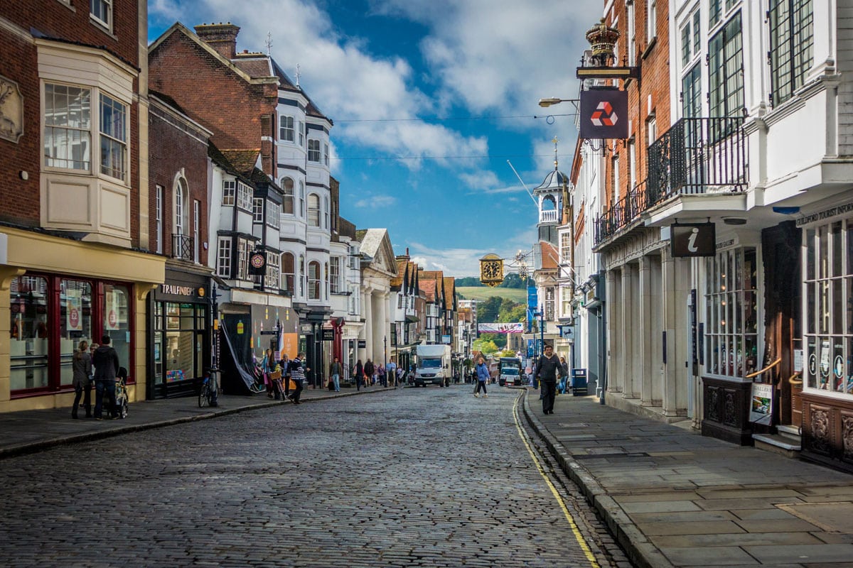 Pedestrians passing shops in Guildford High Street, with a view of the South Downs (hills) in the distance, Surrey, UK.