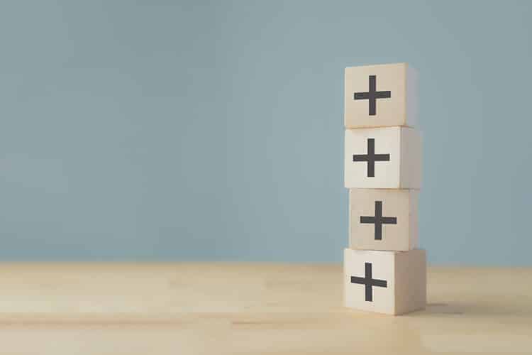 A stack of four wooden cubes each with a plus sign drawn on them Image represents compounding positivity.