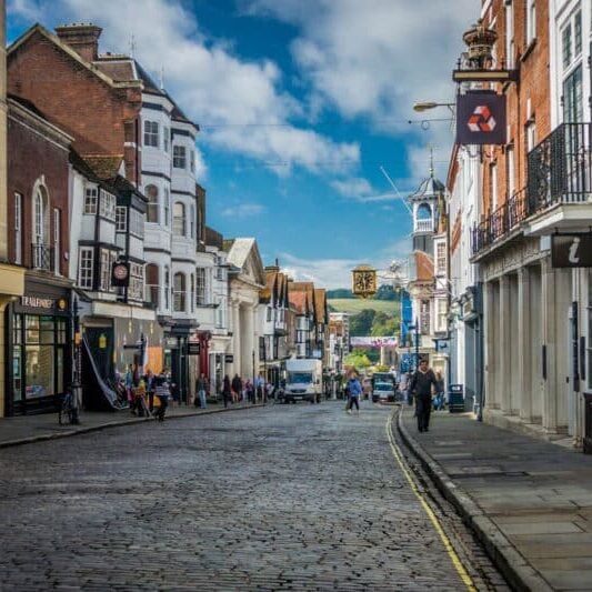 Pedestrians passing shops in Guildford High Street, with a view of the South Downs (hills) in the distance, Surrey, UK.