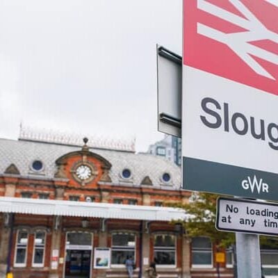 A close up of a sign that says 'Slough' with Slough Train Station in the background