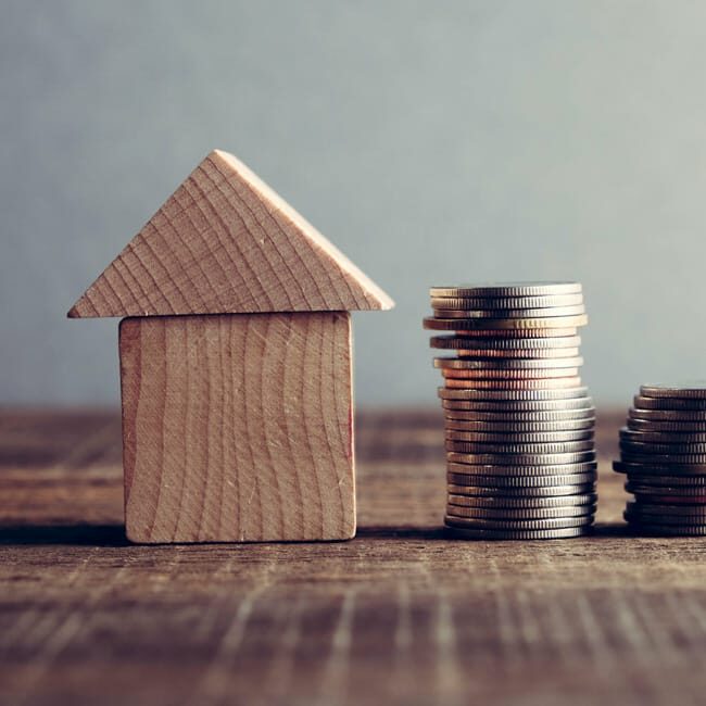 A wooden house sits next to a pile of coins on a table surface. Image represents house value.